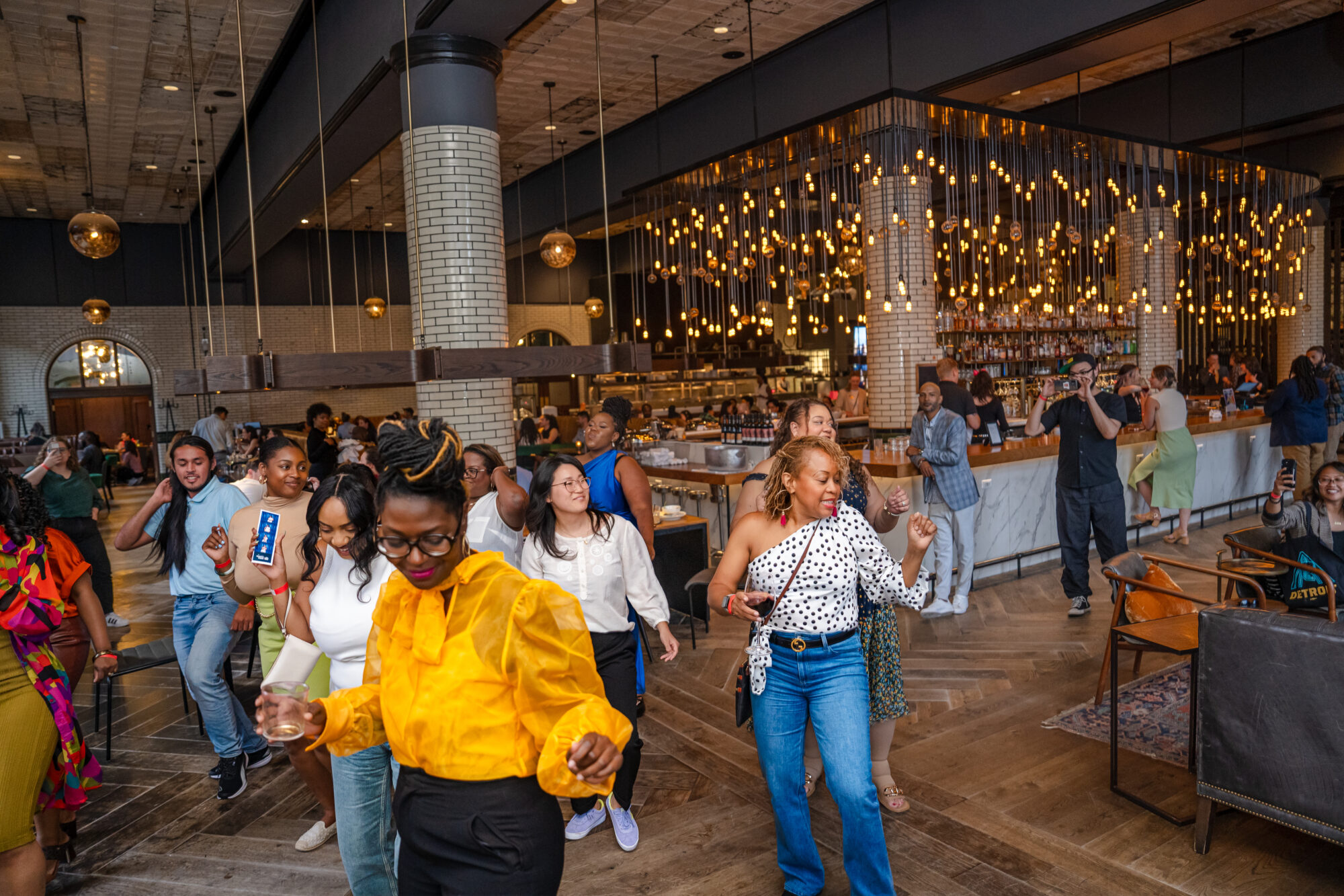 A group of people line dance in a open industrial restaurant with fairy lights hanging in the background over the bar