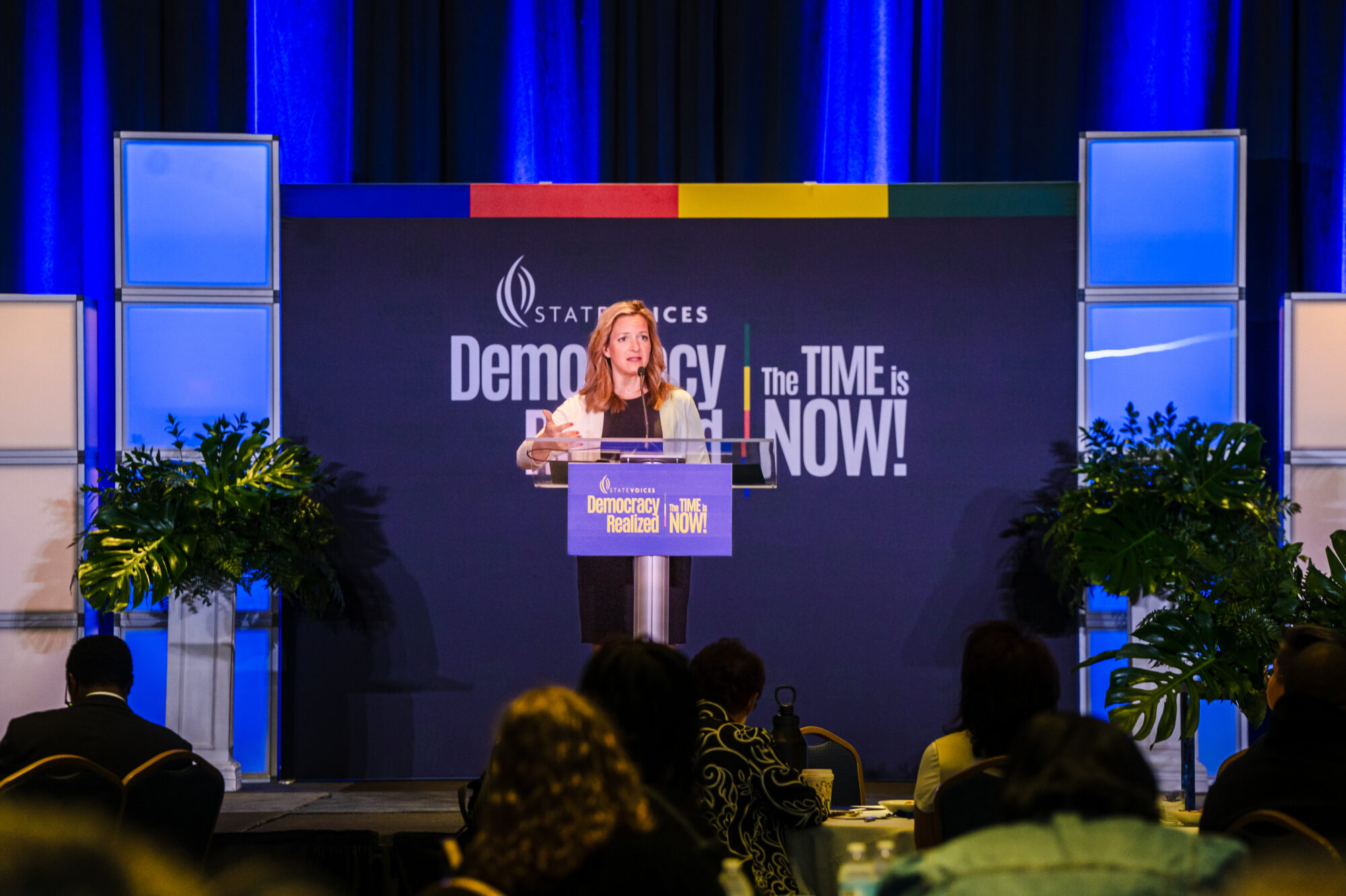 Michigan Secretary of State Jocelyn Benson in a white cardigan and blue dress stands in front of the State Voices Democracy Now banner for her plenary speech. 