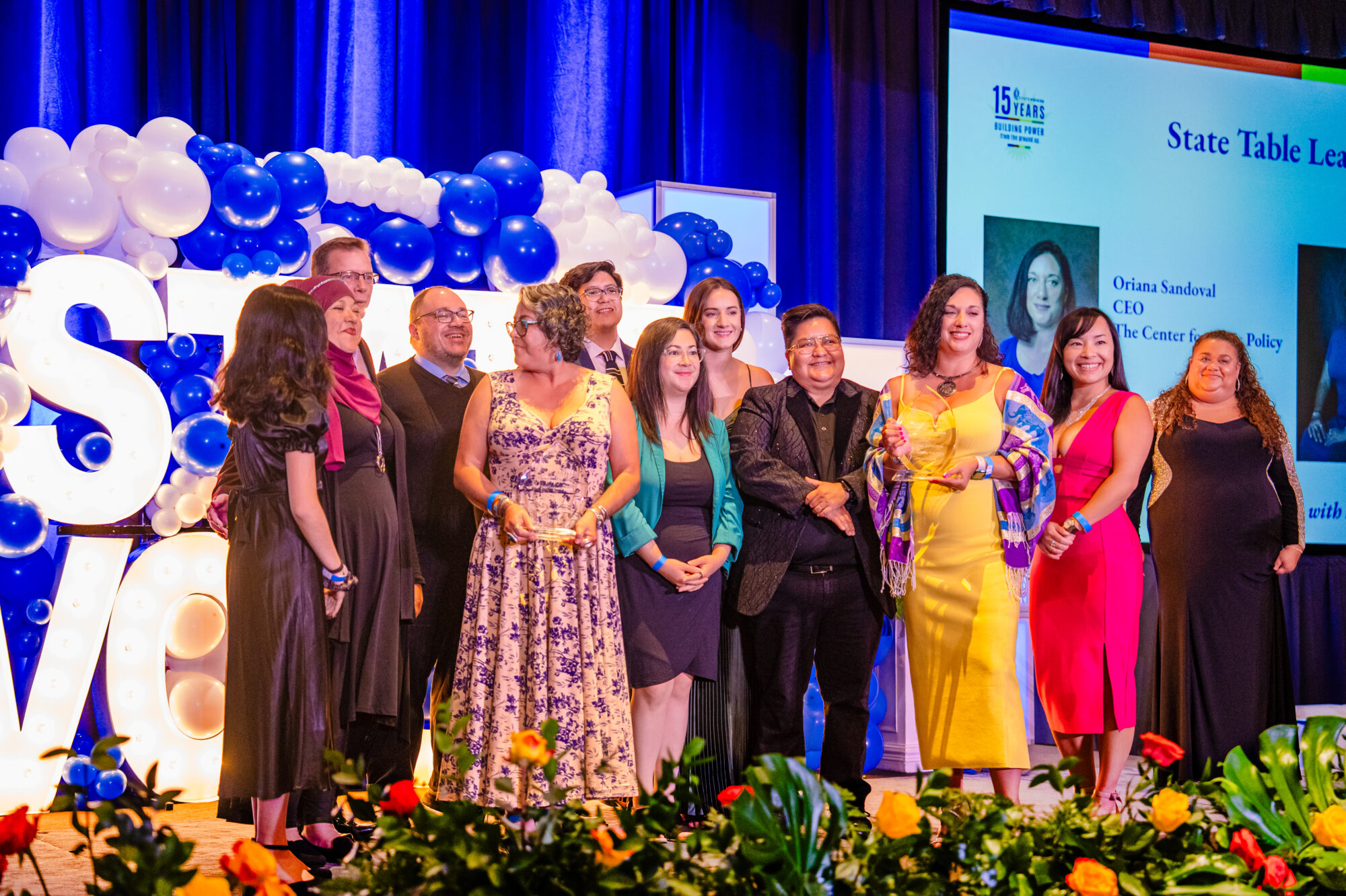 The staff and partners of Center for Civic Policy on stage accepting the State Table Leaders Award. Behind them stands "State Voices" in oversized led lighting with blue and white balloons surrounding the perimeter.